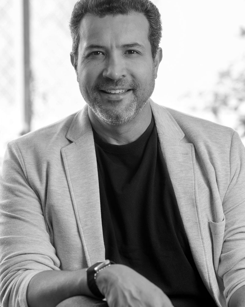Ulrick Noel seated, in a light blazer over a black T-shirt, black-and-white portrait